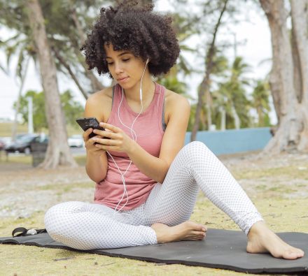 Content fit black woman listening to music with headphones and surfing phone while sitting on mat in park.